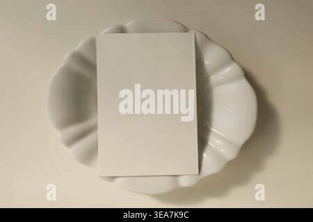 Mockup di biglietti d'auguri vuoti su piatto in ceramica bianca. Sfondo estetico neutro minimo, modello invito vuoto, scena di cancelleria con vista dall'alto, elegante Foto Stock