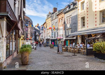 Persone che camminano o si siedono ai tavoli all'aperto su una strada acciottolata costeggiata da architetture storiche a Honfleur in una soleggiata serata estiva Foto Stock