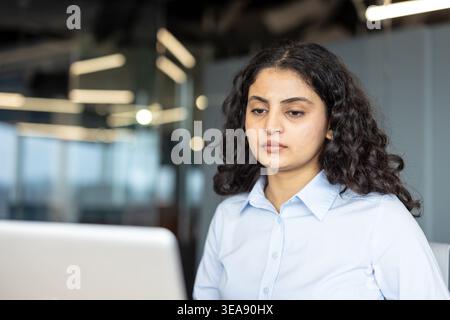 Giovane donna d'affari indiana che si concentra sul suo lavoro, utilizza un notebook in un moderno ambiente aziendale, mostrando professionalità e dedizione ai suoi compiti Foto Stock