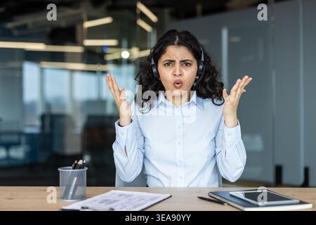 Donna frustrata che indossa una cuffia mentre esprime confusione e incredulità con le mani aperte, ha problemi e stress mentre lavora in un call center in ufficio Foto Stock