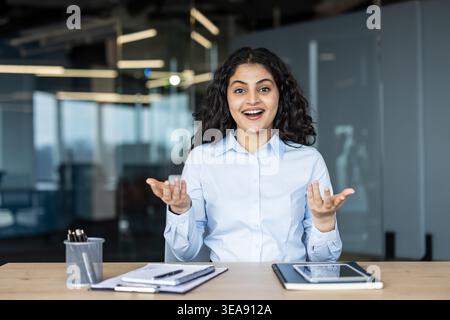 Sorridente giovane donna indiana adulta seduta alla scrivania di un ufficio, gesti eccitanti a mani aperte mentre si partecipa a una videochiamata online o a una discussione di gruppo, esprimendo sorpresa e felicità Foto Stock