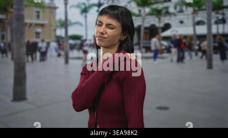 La giovane donna ispanica in maglione Bordeaux afferra e tiene le spalle per il dolore mentre si trova in strada; disagio. Foto Stock