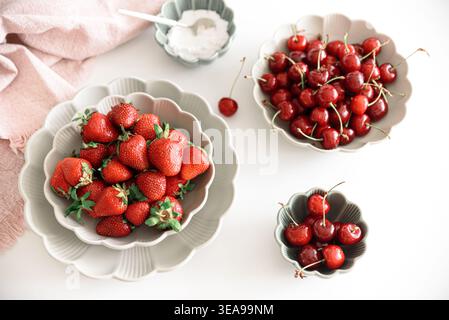 Composizione luminosa e fresca con ciotole di fragole mature e ciliegie rosse, accanto a una piccola ciotola di zucchero su tavola bianca. Foto Stock