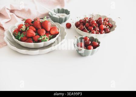 Composizione luminosa e fresca con ciotole di fragole mature e ciliegie rosse, accanto a una piccola ciotola di zucchero su tavola bianca. Foto Stock