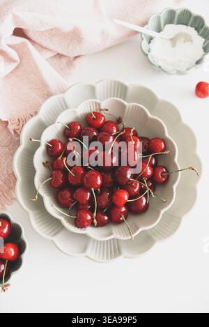 Composizione minimalista con ciliegie rosse fresche con steli disposti in ciotole di ceramica smerlata su un tavolo bianco Foto Stock
