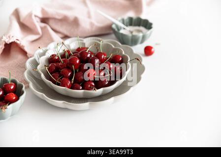 Composizione minimalista con ciliegie rosse fresche con steli disposti in ciotole di ceramica smerlata su un tavolo bianco Foto Stock