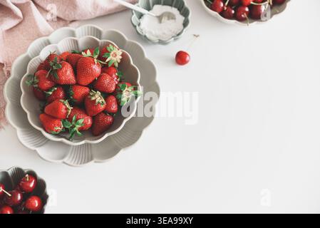 Composizione luminosa e fresca con ciotole di fragole mature e ciliegie rosse, accanto a una piccola ciotola di zucchero su tavola bianca. Foto Stock