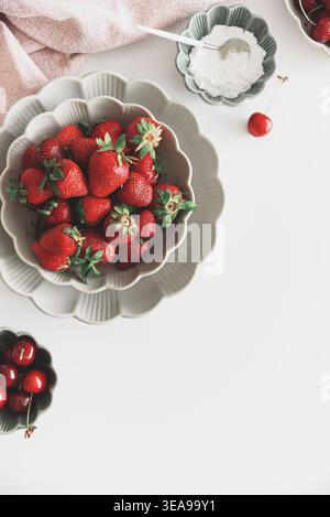Composizione luminosa e fresca con ciotole di fragole mature e ciliegie rosse, accanto a una piccola ciotola di zucchero su tavola bianca. Foto Stock