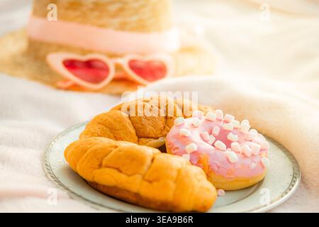 Due croissant e una ciambella giacciono su un piatto blu con un bordo dorato su un asciugamano bianco. Un cappello di paglia e occhiali a forma di cuore si trovano nelle vicinanze, per un picnic in estate Foto Stock