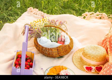 Due croissant e una ciambella giacciono su un piatto blu con bordo dorato su una coperta bianca. Accanto si trova un cappello di paglia e bicchieri a forma di cuore, cestino con daisi Foto Stock