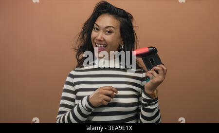 Donna che tiene in mano il controller di gioco e sorride nello studio pesca, con la parte superiore a righe visibile, che mostra la mano tatuata e l'orecchino a cerchio; gioia giocosa. Foto Stock