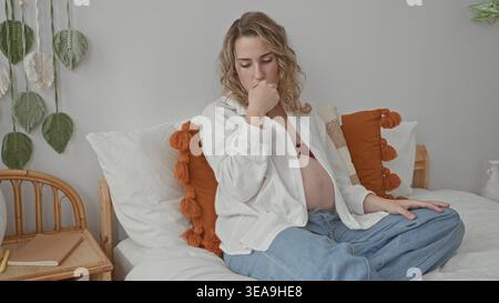 Donna incinta che tocca la bocca con la mano, pancia esposta visibile seduta su un letto in una camera da letto; riposo contemplativo. Foto Stock
