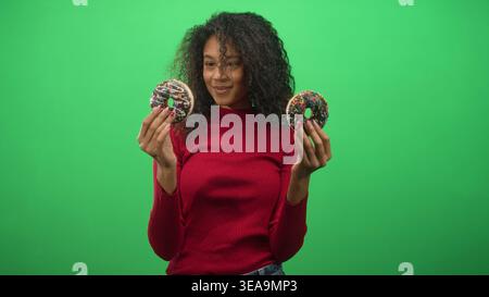 Una ragazza adolescente con capelli ricci tiene due ciambelle cosparse di cioccolato con entrambe le mani contro uno studio di green screen sorridendo; giocosa gioia. Foto Stock
