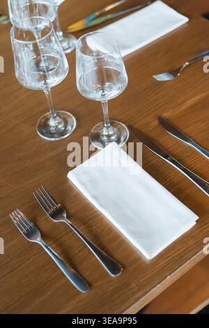 Il tavolo in legno regge un elegante allestimento da pranzo. I bicchieri da vino trasparenti rimangono vuoti sulla superficie. Due forcelle e coltelli poggiano ordinatamente accanto al tovagliolo. Nappa bianca Foto Stock