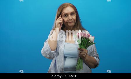 Una donna anziana che tiene in mano tulipani rosa tocca il tempio e punta il dito in studio; riflesso premuroso. Foto Stock