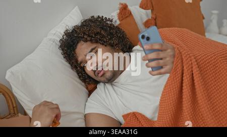 Giovane indiano giacente a letto sotto coperta arancione tiene smartphone, tocca la gola con mano e i principi accanto alla bottiglia di pillola in studio; preoccupazione per la salute. Foto Stock
