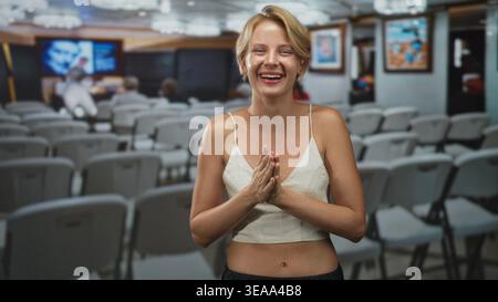 Donna con mani strette che rideva, palme premute vicino al petto in una sala conferenze studio con sedie e schermo; gioia gratitudine. Foto Stock