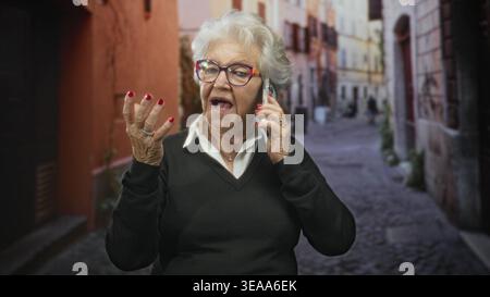 Donna anziana che parla sullo smartphone, punta il dito e i gesti con la mano in una stretta strada acciottolata; connessione sicura. Foto Stock