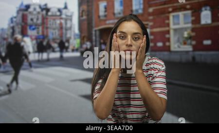Una giovane donna tiene i capelli tra le dita e ispeziona le radici mentre si trova su un attraversamento di strade trafficate della città vicino a edifici in mattoni che indossano un An bianco a righe Foto Stock