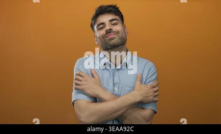 L'uomo attraversa le braccia sul petto abbracciandosi con gli occhi chiusi e un sorriso nello studio arancione; serenità. Foto Stock