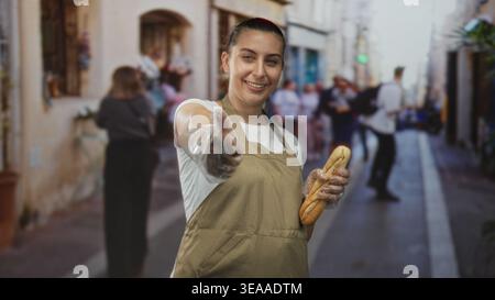 Donna in grembiule che tiene in mano la baguette con i guanti di plastica e che dà i pollici in strada; gioiosa ospitalità. Foto Stock