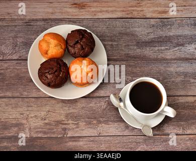 Una tazza di caffè nero e un piatto con quattro cupcake di cioccolato fresco e vaniglia su un tavolo di legno. Vista dall'alto, disposizione piatta. Foto Stock