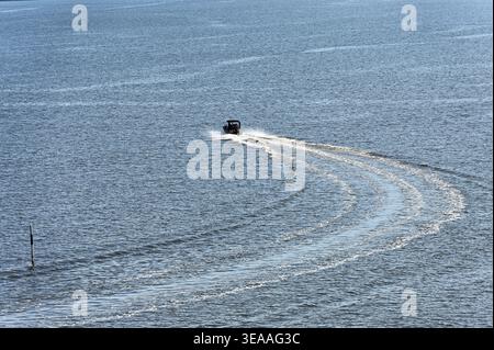 Una piccola barca a motore si muove in mare aperto lasciando dietro di sé un'ampia scia curva, creando motivi eleganti sulla superficie calma e trasportando il movimento Foto Stock