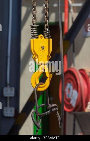 il gancio per gru industriale giallo brillante con un fermo di sicurezza è sospeso da catene in acciaio pesante e da un cavo metallico in un'officina all'aperto illuminata dal sole. Foto Stock