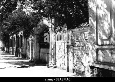 Slavyansk, 1970 anni Vista dettagliata a livello della strada che si concentra direttamente su un arco storico in mattoni e una spessa parete di confine vicino ai grandi magazzini in Karl Marx Street (ora Tsentralna Street). L'intensa luce del sole proietta ombre nette dal fogliame circostante, enfatizzando l'intonaco sbriciolato e il decadimento architettonico. Questa accattivante fotografia d'archivio in bianco e nero funge da monumentale testimonianza del patrimonio urbano della città anteguerra, immortalando splendidamente le tranquille e senza tempo strade del tranquillo Donbas. Foto Stock