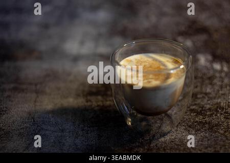Vista laterale del caffè sporco in vetro a doppia parete su un tavolo in cemento scuro. Loft industriale in stile caffetteria con una suggestiva illuminazione laterale. Foto Stock
