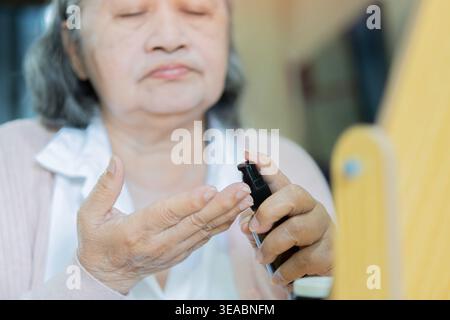 Donna asiatica anziana che si prende cura della sua pelle, applicando siero anti-invecchiamento davanti a uno specchio a casa. Messa a fuoco selettiva. Concetto di cura della pelle per anziani. Foto Stock