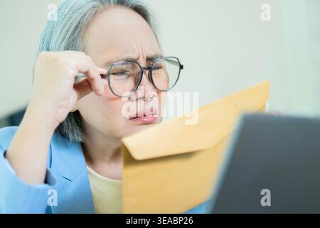 La vista laterale di una donna matura con gli occhiali cerca di leggere una busta marrone, usando un laptop a casa, avendo problemi di vista. Soffre di problemi agli occhi. Foto Stock