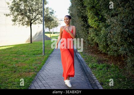 Elegante giovane signora vestita di arancio che esplora i lussureggianti sentieri del parco durante il giorno Foto Stock