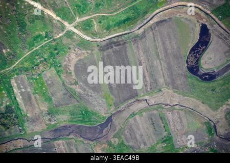 Vista aerea di un piccolo fiume serpeggiante che scorre attraverso terreni agricoli coltivati con motivi di terreno scuri, prati primaverili e strade di campagna che creano una geometria naturale astratta. Foto Stock