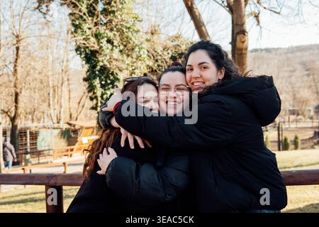 Tre sorelle caucasiche si abbracciano e sorridono alla macchina fotografica in un parco zoo con alberi e recinzioni in sottofondo durante il giorno. Famiglia, Relationshi Foto Stock