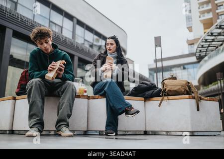 Amici delle scuole superiori che mangiano panini e bevande seduti sulle panchine urbane all'aperto vicino ai negozi Foto Stock