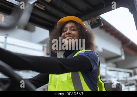 Ritratto di una lavoratrice sorridente che indossa indumenti protettivi, un elmetto giallo e un giubbotto di sicurezza, che guida un carrello elevatore a forche in un magazzino. Foto Stock