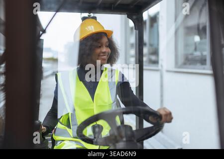 Ritratto di una lavoratrice sorridente che indossa indumenti protettivi, un elmetto giallo e un giubbotto di sicurezza, che guida un carrello elevatore a forche in un magazzino. Foto Stock