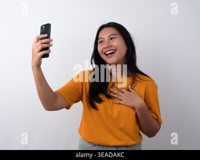 Felice consapevole grato giovane donna asiatica sorridente mentre tiene il telefono con le mani sul petto su sfondo bianco isolato. Foto Stock