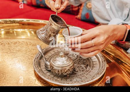 Versare a mano il tradizionale caffè turco da una crosta di metallo ornata in una piccola tazza su un vassoio d'argento e oro inciso Foto Stock