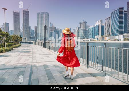 Giovane donna in abito rosso con zaino e cappello che cammina lungo una passeggiata sul lungomare con il moderno skyline della città di Dubai Foto Stock