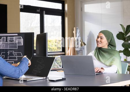Team diversificato che collabora in ufficio, donna in hijab verde che utilizza notebook, monitor CAD, spazio di copia Foto Stock