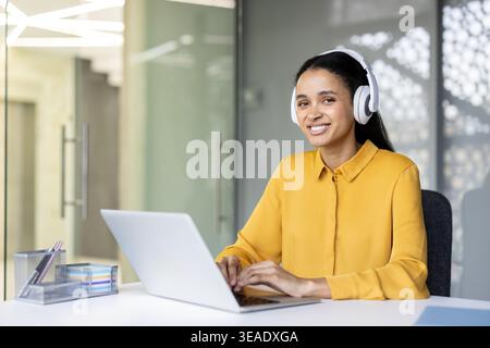 Giovane donna d'affari che indossa cuffie wireless bianche e una camicia gialla che scrive su un laptop, sorridendo verso la fotocamera mentre lavora a una scrivania in un ambiente d'ufficio contemporaneo Foto Stock