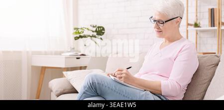 Una donna anziana si impegna a scrivere mentre si siede comodamente sul divano in una camera luminosa Foto Stock