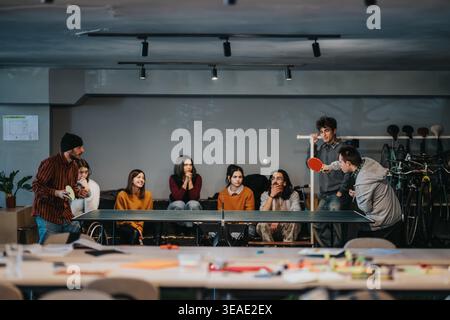 Gli studenti giocano a ping pong in una sala informale con gli amici che guardano e socializzano Foto Stock