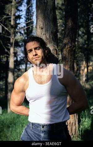 Un uomo robusto e atletico con capelli scuri e barba si trova in una foresta dondolata dal sole, il suo fisico muscoloso evidenziato dalla canotta bianca e dai jeans Foto Stock