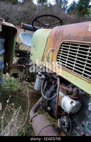 Primo piano del vecchio motore e del pannello della carrozzeria del trattore arrugginito con volante in un campo rurale erboso con alberi e arbusti sullo sfondo. Foto Stock