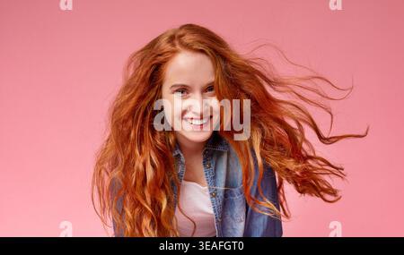 La giovane donna con lunghi capelli rossi sorride gioiosamente su uno sfondo rosa mentre posa giocosamente Foto Stock