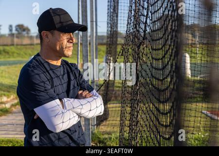 Uomo maturo in piedi a sinistra, indossa un cappellino da baseball con reti e pali sul campo, copia spazio Foto Stock