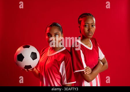 Due donne afroamericane in posa in studio con maglie da calcio rosse-bianche abbinate che tengono la palla Foto Stock